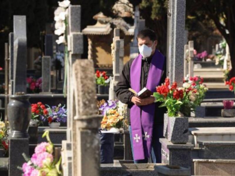 Un sacerdote con máscara y sotana celebra una misa en un cementerio con flores y lápidas alrededor.