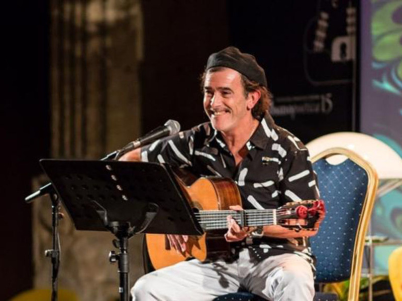 Un músico en un concierto, tocando la guitarra y sonriendo, con un fondo de luces y una pantalla con un diseño de ondas azules y verdes.