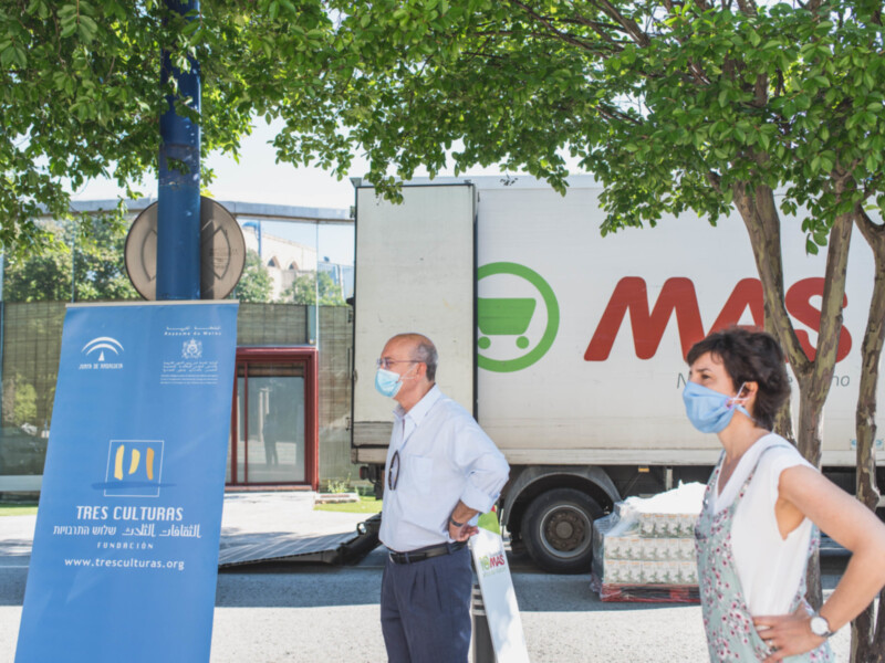 Dos personas con mascarillas frente a un camión de la Fundación Tres Culturas y el logo de MAS, en un entorno verde y soleado.