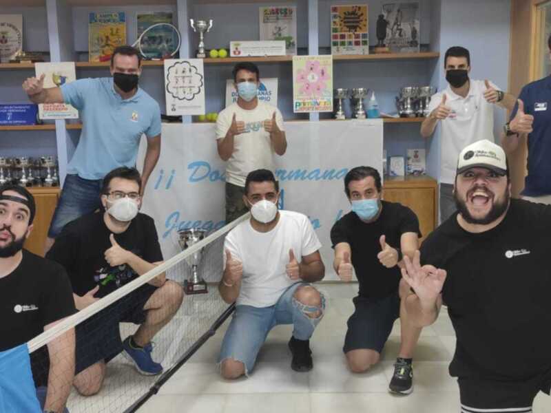 Un grupo de personas con máscaras faciales sonríen y dan el 'OK' con las manos, posando en una oficina con estantes de trofeos y carteles de eventos deportivos.