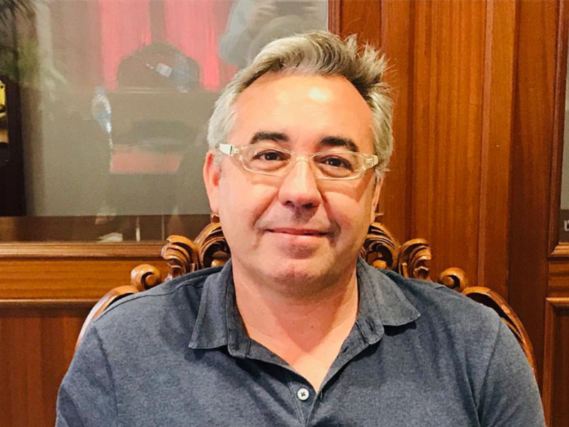 Hombre con camisa azul y gafas, sentado en un sillón de madera con fondo de pared de madera y puerta trasera.