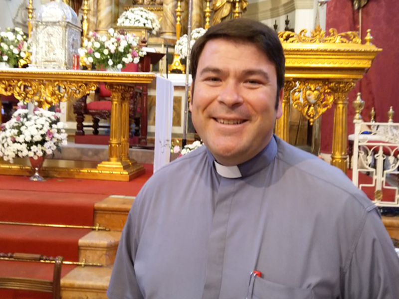Retrato de un sacerdote sonriente en una iglesia con detalles dorados y flores blancas.