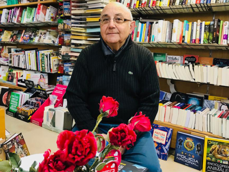 Un hombre en una librería con flores rojas y libros de diferentes títulos en el fondo.