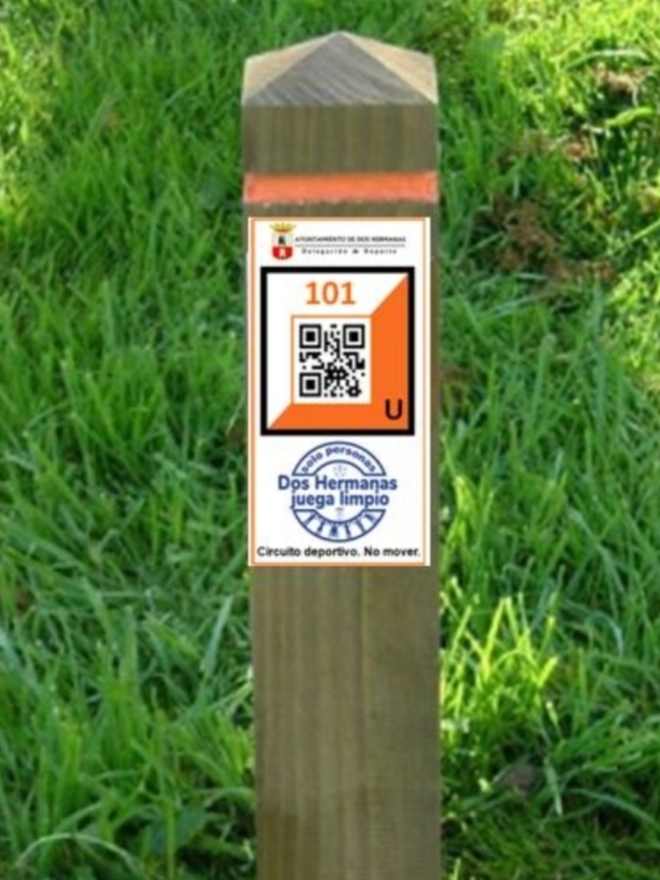 Un poste de madera con un letrero que indica 'Circuito deportivo. No mover.' y un código QR en el fondo de un césped verde.