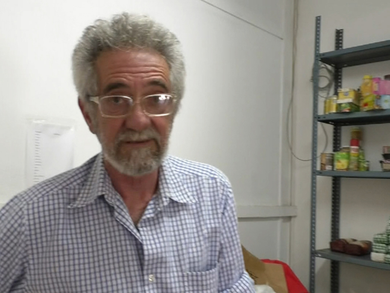 Un hombre con barba y gafas, posiblemente en un almacén, frente a una estantería con productos en el fondo.