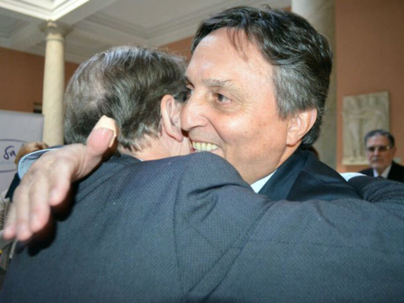 Dos hombres saludándose en un evento formal, uno sonríe ampliamente mientras el otro le da un abrazo. En el fondo, se puede ver una columna y una pintura en el muro. La imagen sugiere un ambiente de amistad o celebración.