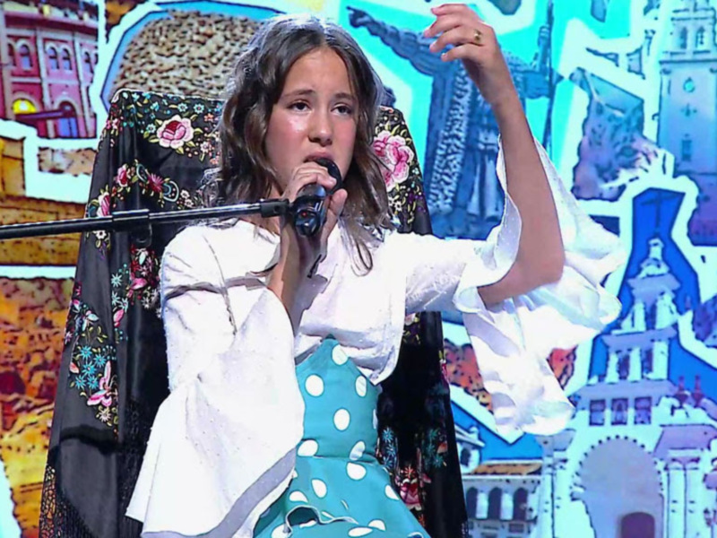 Una joven cantante en un escenario, con un vestido blanco y azul con lunares, frente a un fondo con imágenes de edificios históricos y arquitectura.