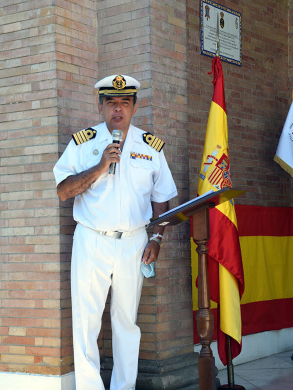 Un oficial naval en uniforme blanco habla en un micrófono, con una bandera española a su lado y un letrero en la pared de fondo.