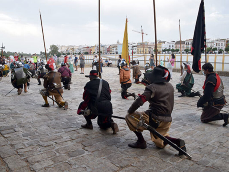 Recreación histórica de una batalla medieval con soldados en armadura y banderas en un escenario urbano.