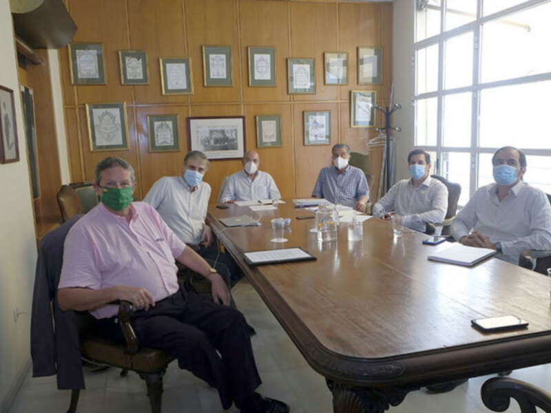 Reunión de trabajo en una sala de conferencias con hombres sentados alrededor de una mesa, todos con mascarillas y documentos en el centro.