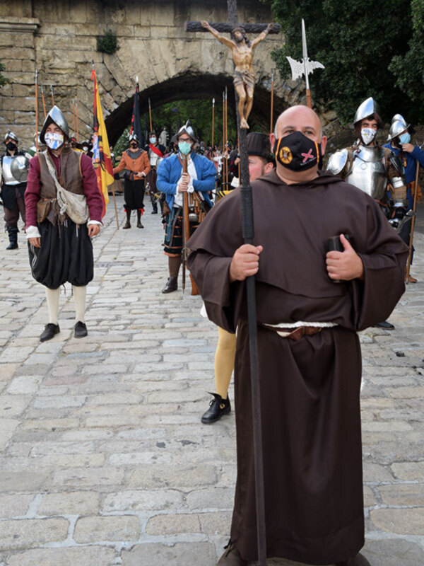 Procesión histórica con monje portando una cruz y personajes vestidos con trajes medievales, con máscaras faciales en un entorno urbano.