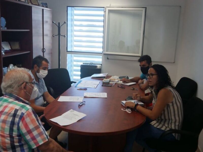 Un grupo de personas en una reunión de trabajo, todos con mascarillas, sentados alrededor de una mesa con documentos y materiales de oficina. La sala tiene una puerta con persianas abiertas y una pizarra en la pared. El ambiente es formal y profesional.