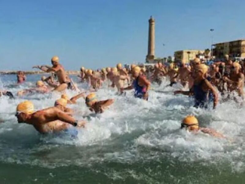 Competidores en una carrera de natación abierta en el mar, con un faro al fondo bajo un cielo despejado.