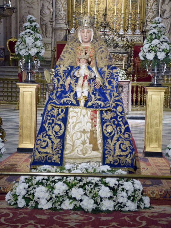 Virgen de la Cinta en su trono ornado con flores blancas, vestida con un manto azul y dorado en un interior de iglesia con decoración barroca.