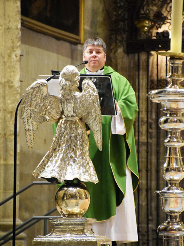 Un sacerdote vestido con un hábito verde y blanco, posando junto a una estatua de ángel dorada en una iglesia.