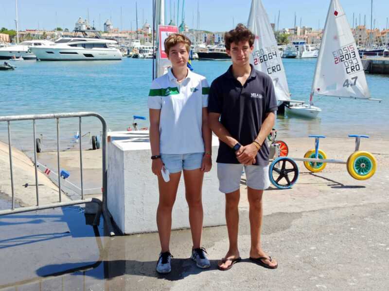 Dos jóvenes posan en un puerto con barcos de vela en el fondo.