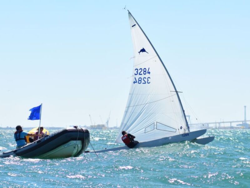 Competición de vela en el mar, con barco a vela y remo en primer plano, bajo un cielo despejado.