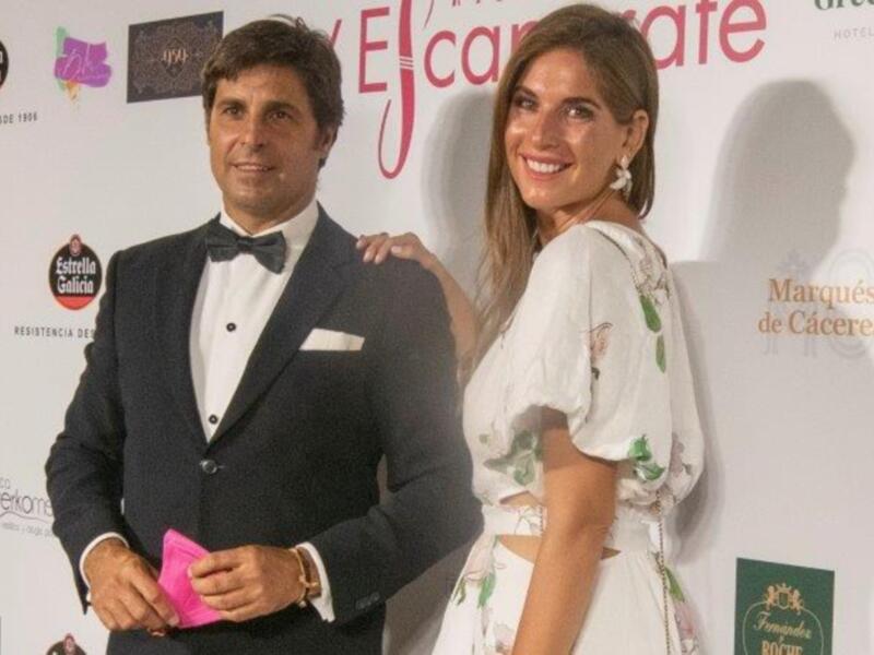 Una pareja elegante posa juntos en un evento formal, con el hombre en traje y la mujer en vestido blanco bordado.