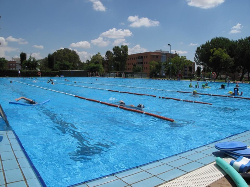 Piscina pública con personas nadando y disfrutando del día bajo un cielo azul con algunas nubes dispersas.