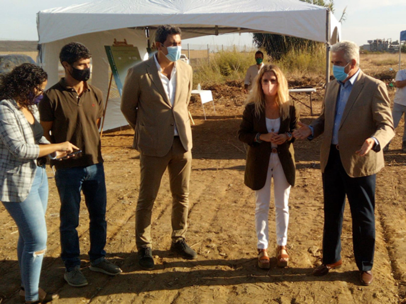 Un grupo de personas con máscaras se reúne bajo una carpa en un área de construcción o desarrollo, posiblemente discutiendo sobre un proyecto en curso.