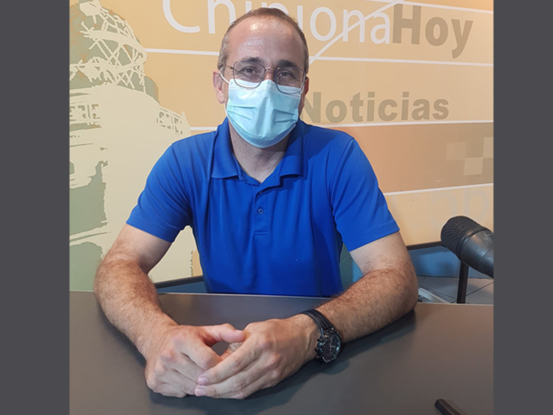 Un hombre en una entrevista, con una camiseta azul y una máscara médica, frente a un fondo con el logo de una empresa de noticias.
