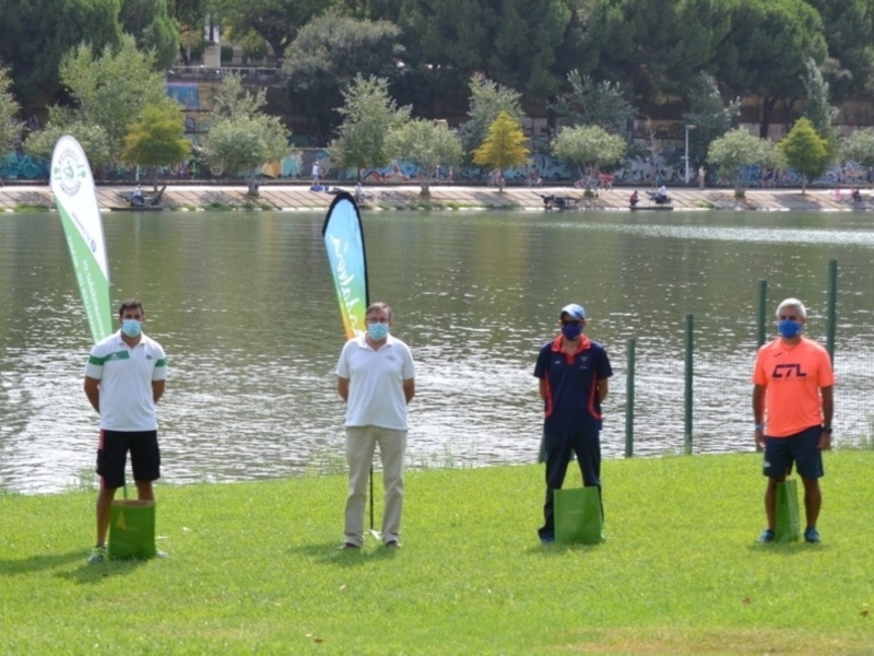 Gente de pie en el parque con pancartas y bolsas de compras en la orilla del lago, con árboles y grafitis en el fondo.