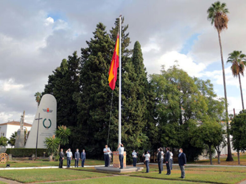Ceremonia de izado de la bandera en un entorno conmemorativo y árboles al fondo.