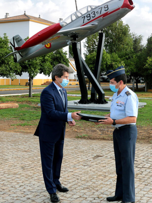 Dos personas en uniforme militar, uno de ellos con una máscara médica, sostienen un objeto en un entorno verde con un avión militar en el fondo.