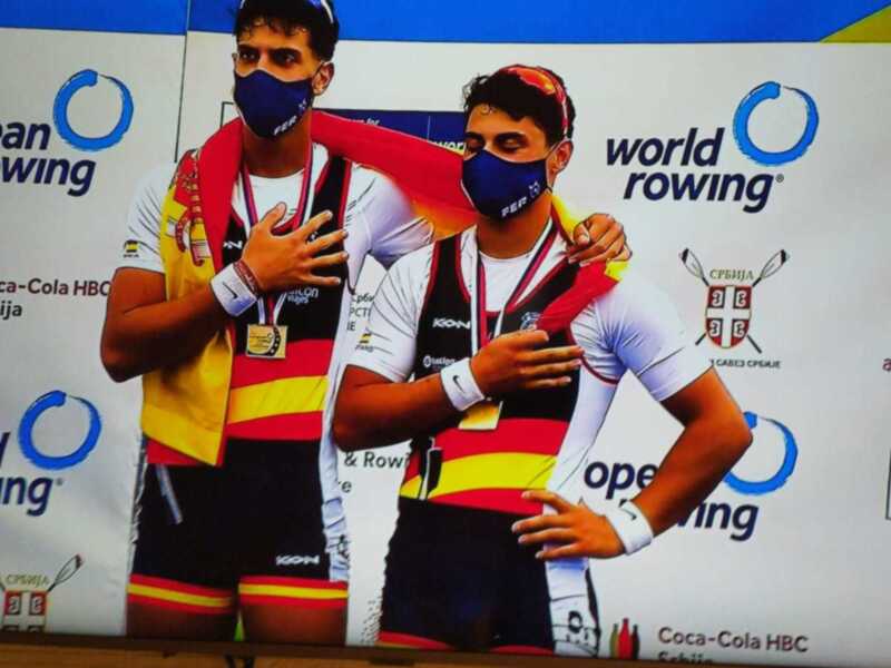 Dos atletas posan en un podio con medallas, en el contexto del Campeonato Europeo de Remo. El fondo muestra logos de patrocinadores como Coca-Cola y World Rowing.