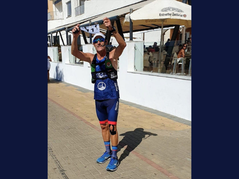 Un atleta en traje de triatlón posa con orgullo tras completar una carrera de triatlón en una playa con sombrillas y edificios blancos al fondo.