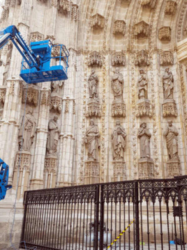 Trabajos de mantenimiento en la fachada de una catedral gótica con detalles escultóricos y una plataforma elevadora azul en primer plano.