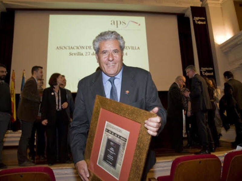 Premio recibido por un hombre en un evento formal, con una pantalla en el fondo que dice 'ASOCIACIÓN DE SEVILLA'.