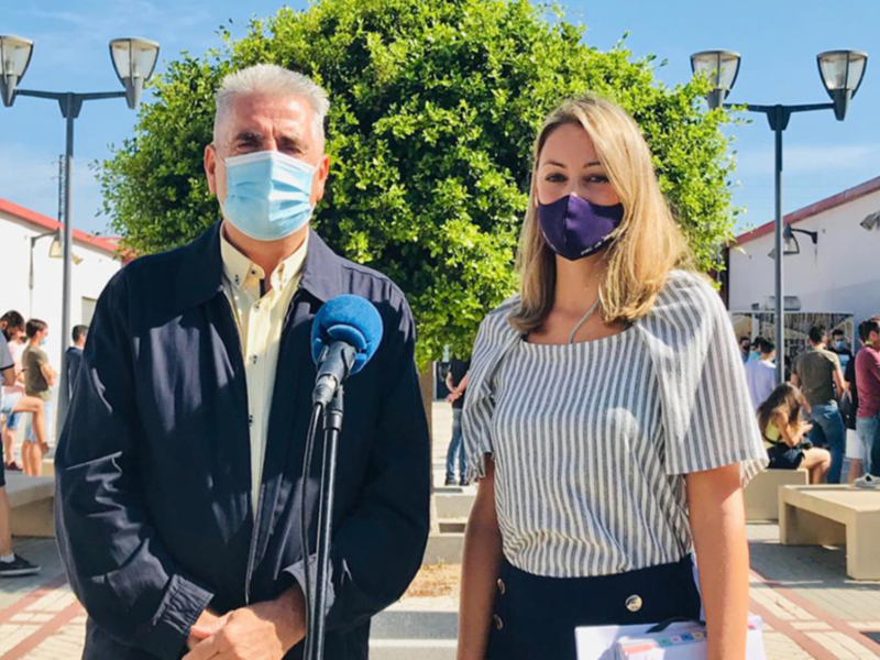 Persona con micrófono y otra con máscara facial en evento al aire libre, con árboles y personas en segundo plano.