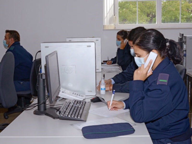Persona trabajando en un escritorio con computadora, hablando por teléfono y usando mascarilla, en un ambiente de oficina con otras personas en el fondo.