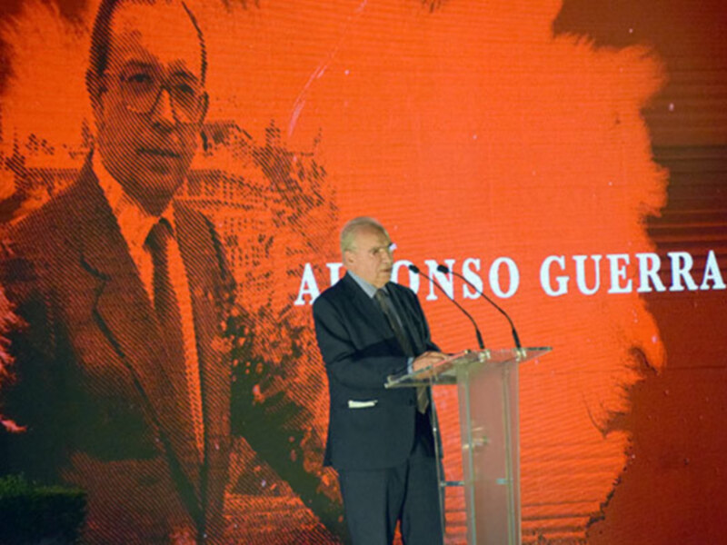 Un hombre en traje habla desde un podio en un evento formal con un fondo rojo y una imagen de un rostro en la pantalla.