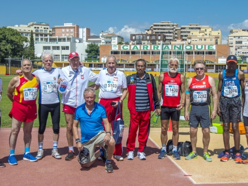 Equipo de atletas senior posando en una pista de atletismo, con banderas nacionales en sus uniformes. Fondo: edificios y cielo azul claro.
