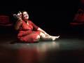 Una mujer en un vestido rojo se sienta en el suelo de una escena teatral, rodeada por sillas rojas. La iluminación es dramática con un foco en su rostro y piernas, creando una atmósfera de tensión.