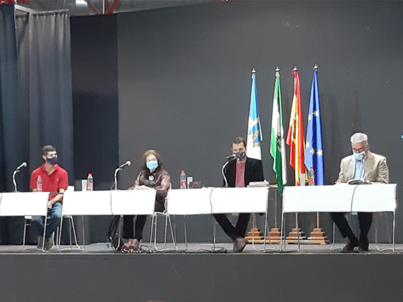 Una conferencia o evento público con personas en una mesa de conferencia, todos con mascarillas, frente a una pantalla y banderas. La escena sugiere una reunión formal o política.
