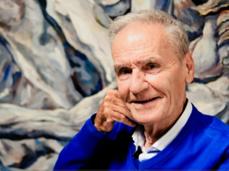 Retrato de un hombre mayor sonriente, vestido con un suéter azul, frente a un fondo abstracto de colores.