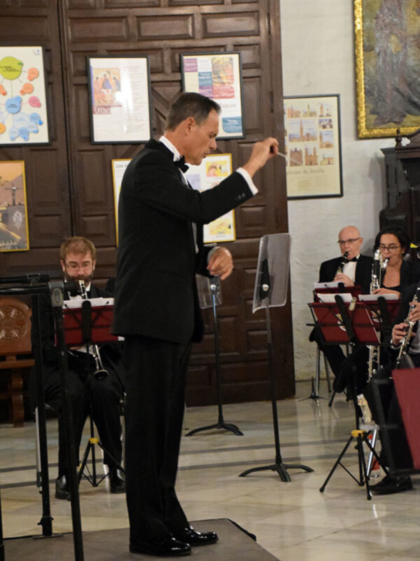 Concierto de música clásica en un entorno acogedor con un director de orquesta en el centro y músicos tocando instrumentos de viento.