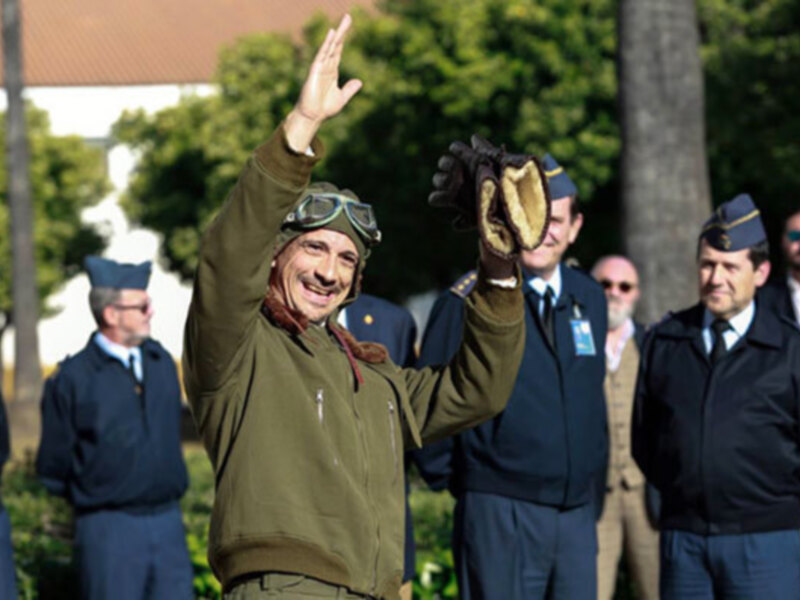 Un hombre en uniforme militar saluda con la mano mientras otros uniformados observan en un entorno al aire libre.