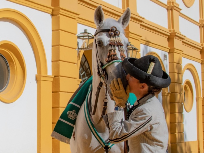 Hombre en traje tradicional besando a un caballo blanco adornado, frente a una fachada amarilla y blanca.