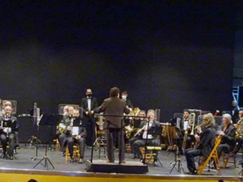 Orquesta sinfónica en un concierto, con músicos tocan instrumentos de viento y percusión en una escena dramática con telón de fondo negro y luces de escenario.