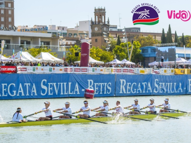 Equipo femenino remando en una regata en Sevilla, con espectadores y banderolas de patrocinio al fondo.