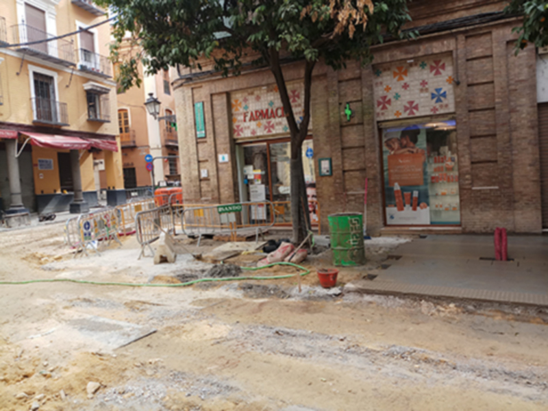 Trabajos de construcción en una calle urbana, con tienda de farmacia y edificios de apartamentos al fondo.