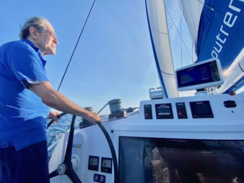 Un marinero maneja el timón de un velero mientras navega bajo un cielo azul claro.
