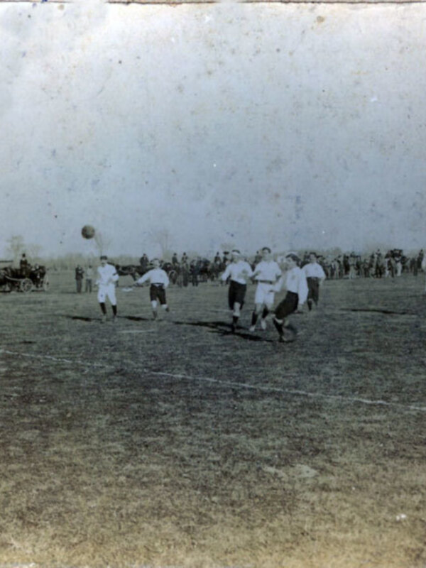 Historic football match on a grassy field, with players in vintage attire and a large crowd in the background.