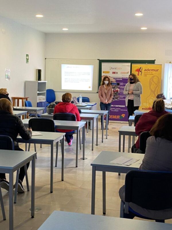 Una clase en un salón de actos con estudiantes sentados en mesas y dos personas en el centro, una de ellas con una presentación en la pantalla.