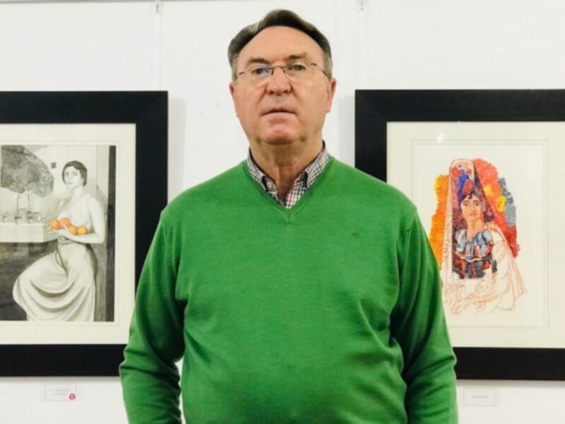 Un hombre en una sala de exposición de arte, posando frente a dos cuadros, uno de ellos es una ilustración de una mujer con un vestido rojo y otro es una escena con una figura femenina en blanco y negro.