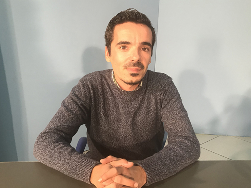 Un hombre con una camisa de manga larga y un suéter gris, sentado en una silla frente a una pared blanca.